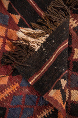 Detalle de alfombra doblada que muestra el reverso, los flecos y los patrones geométricos en tonos rojo, negro, beige y azul oscuro.