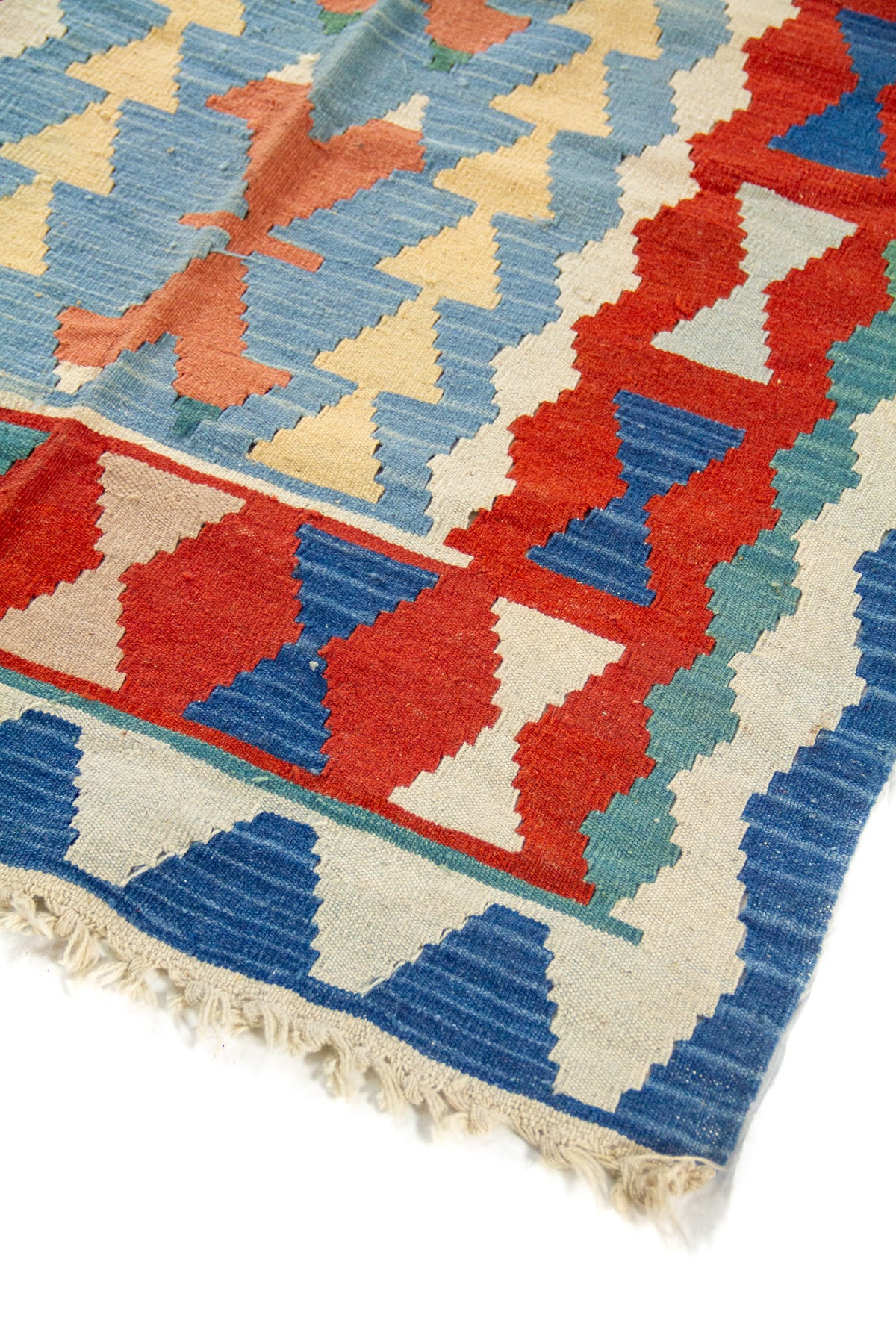 Detalle de esquina de alfombra geométrica con borde azul, figuras escalonadas en rojo y celeste, y flecos en tono beige.
