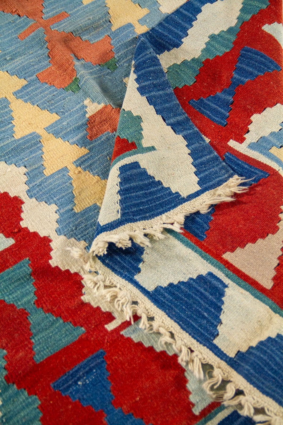 Detalle de alfombra artesanal doblada con motivos geométricos en azul, beige y rojo, mostrando textura y acabado tejido.