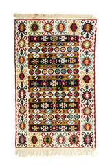 Alfombra rectangular tipo kilim en tonos blanco suave, rojo, verde, celeste y negro, con diseño geométrico multicolor y flecos en ambos extremos.