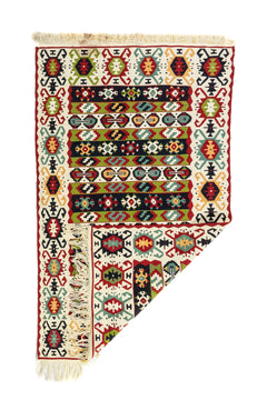 Alfombra geométrica artesanal con una esquina doblada, en colores, blanco, rojo, verde y negro, que muestra su textura y flexibilidad.