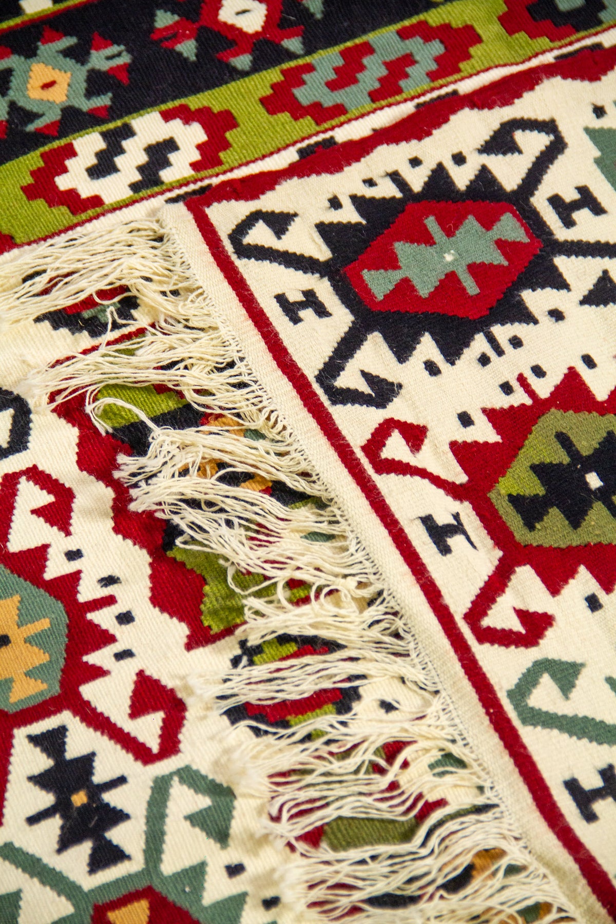 Detalle de alfombra doblada con flecos visibles y diseño geométrico tribal en colores blanco, rojo, verde, celeste y negro.