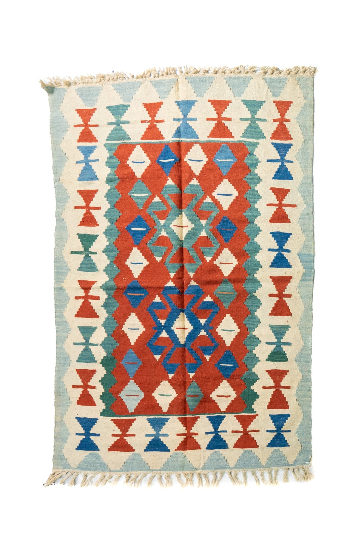 Alfombra rectangular artesanal en tonos celeste, beige, rojo  y azul, con diseño geométrico simétrico y flecos en ambos extremos.
