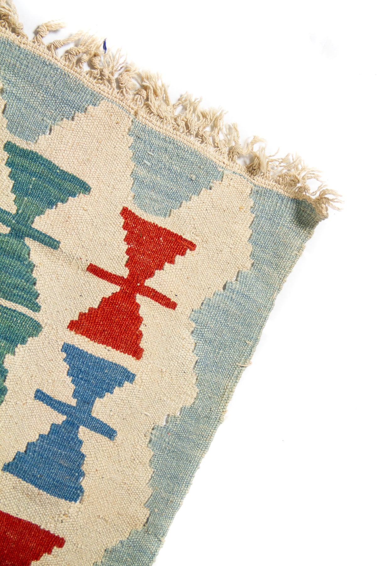Acercamiento al borde superior de alfombra artesanal con patrones geométricos repetitivos en rojo, azul sobre fondo beige.