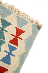 Acercamiento al borde superior de alfombra artesanal con patrones geométricos repetitivos en rojo, azul sobre fondo beige.
