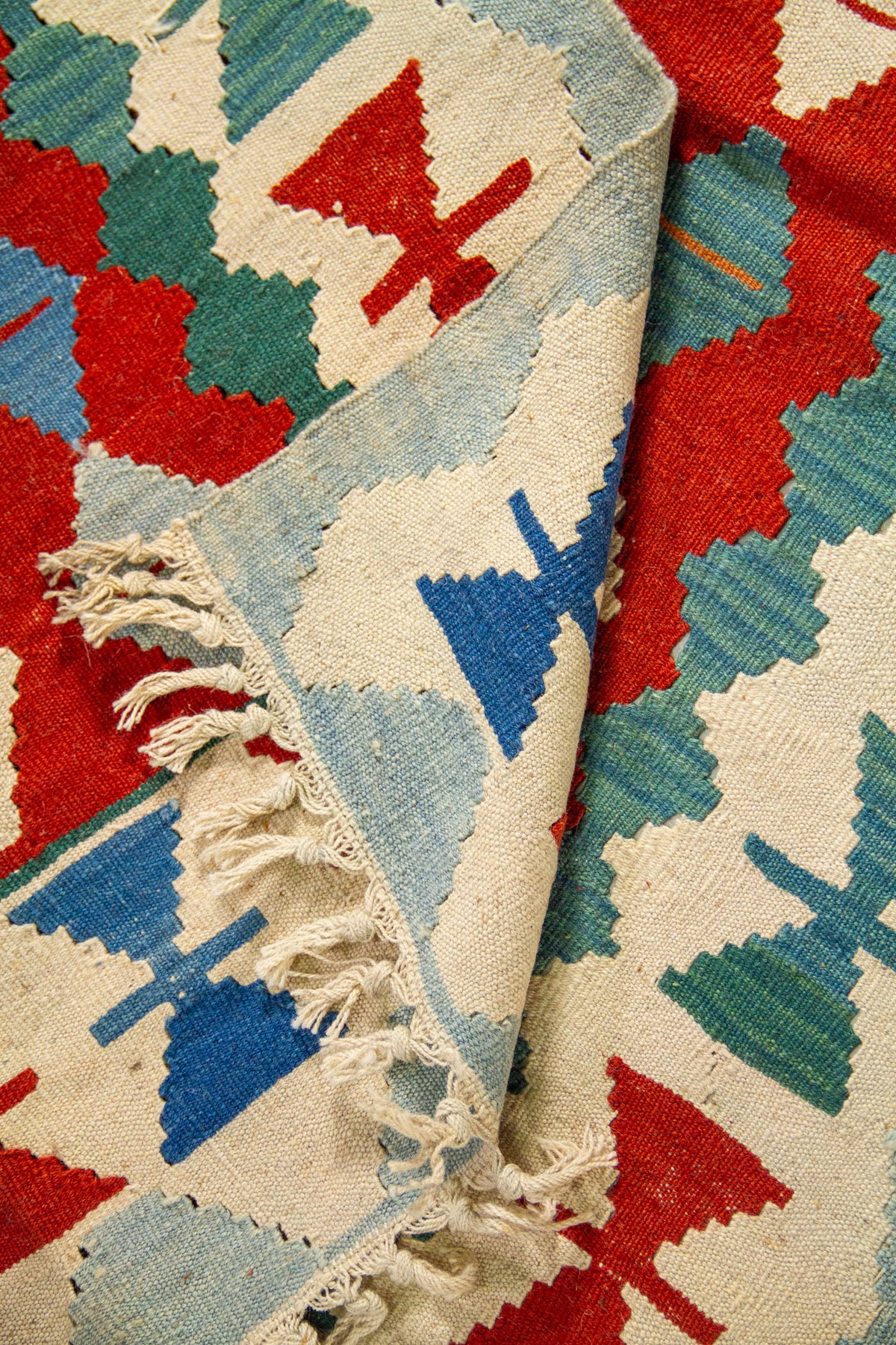 Detalle de alfombra doblada que muestra el reverso, los flecos y los motivos geométricos en tonos celeste, beige, rojo y azul intenso