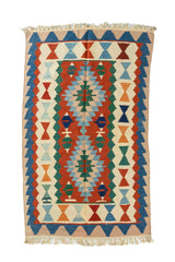 Alfombra rectangular artesanal en tonos beige, azul, rojo, verde, con diseño geométrico simétrico y flecos en ambos extremos.