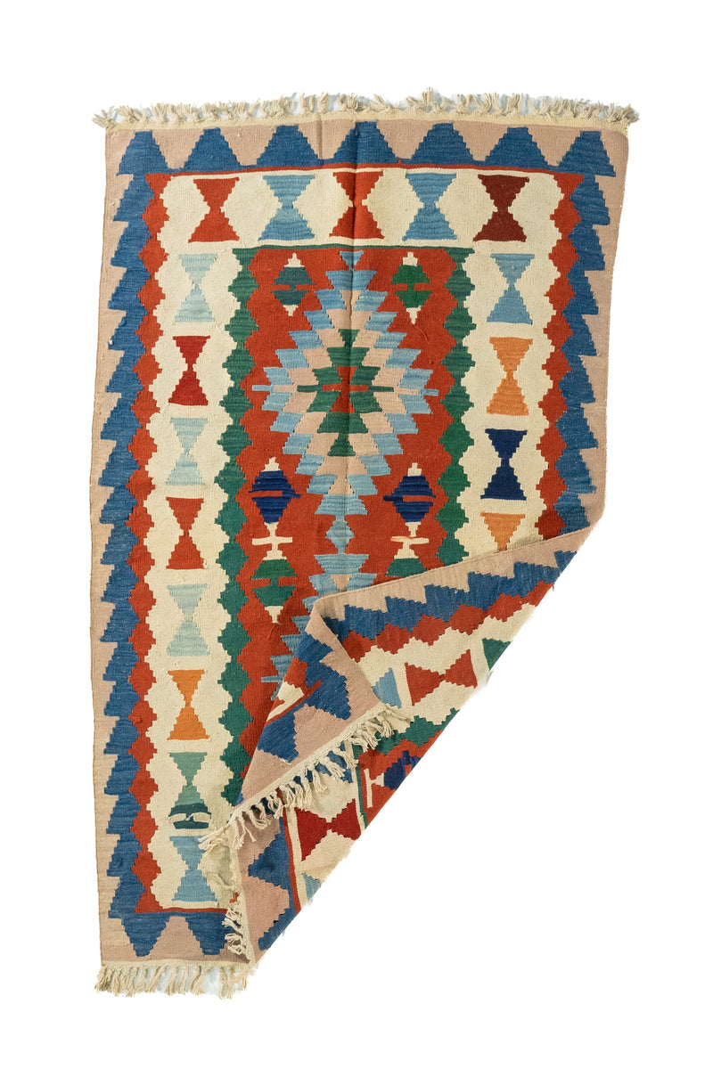 Alfombra geométrica tejida con una esquina doblada, en colores beige, azul, rojo y verde, que muestra su flexibilidad y acabado artesanal.