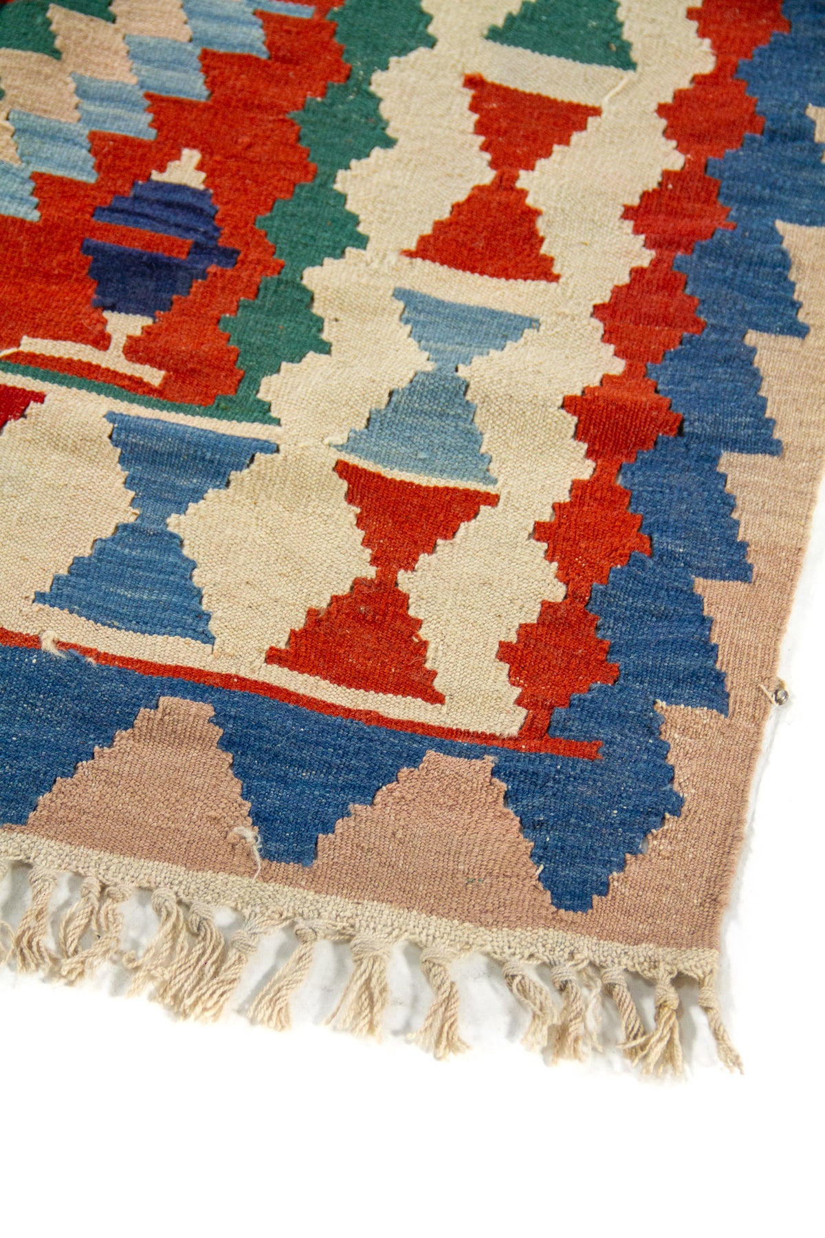 Detalle de esquina de alfombra con borde azul, figuras geométricas en rojo y celeste sobre fondo beige, y flecos tejidos en tono natural.