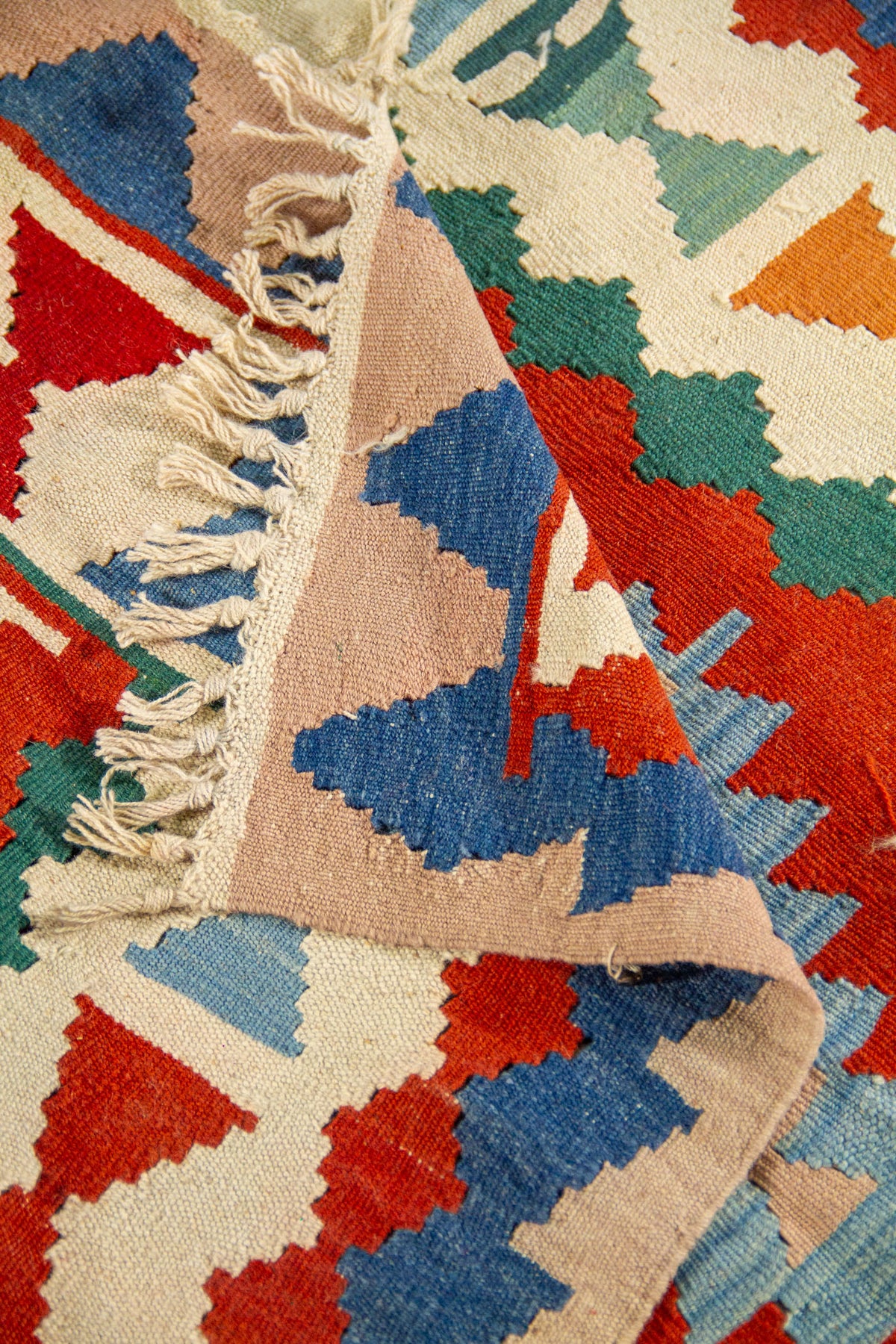 Detalle de alfombra doblada que muestra el reverso, los flecos y los patrones geométricos en tonos beige, azul, rojo y verde 