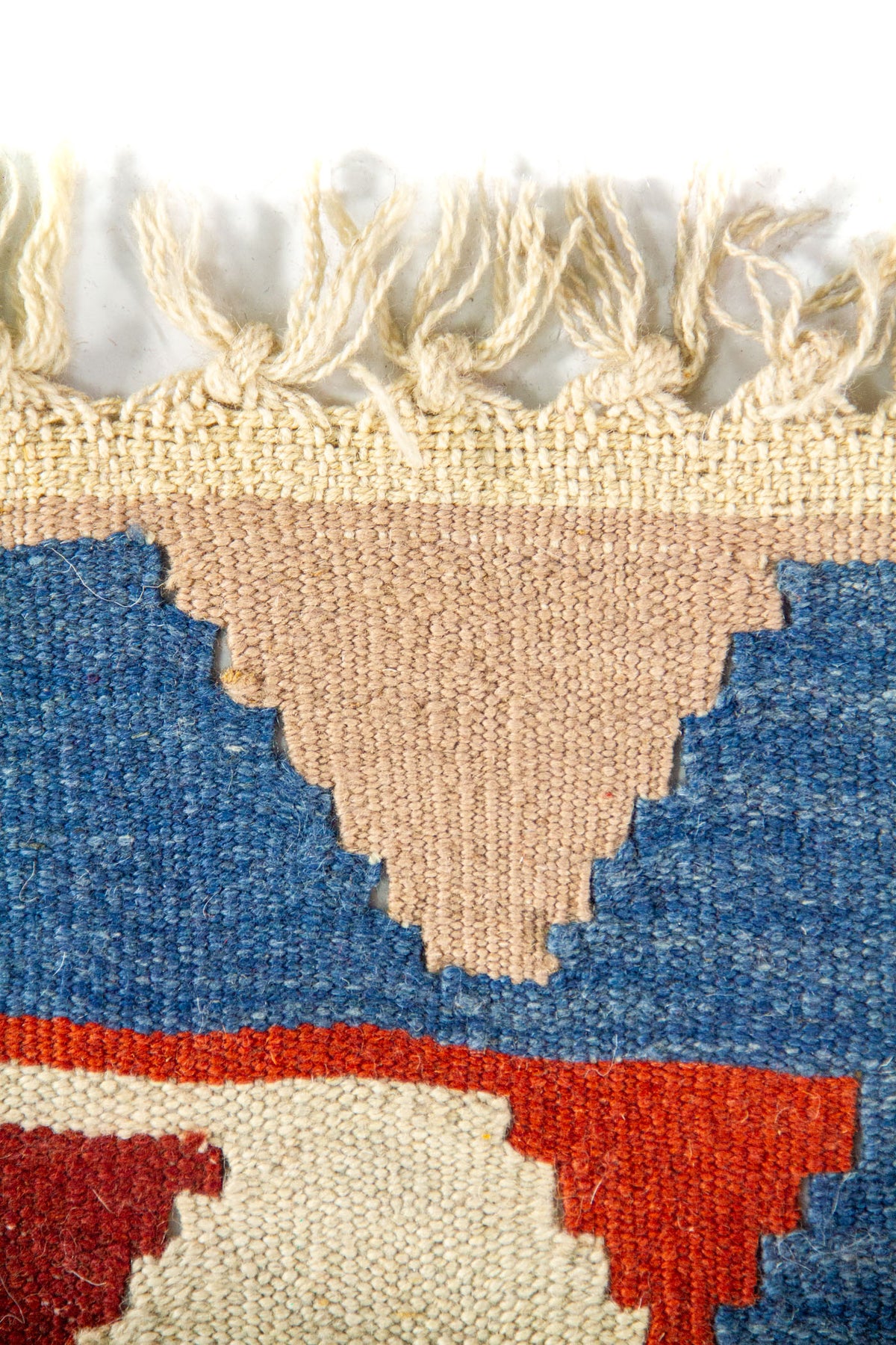 Detalle superior de alfombra tejida con flecos en tono natural y borde geométrico en beige, azul y rojo.