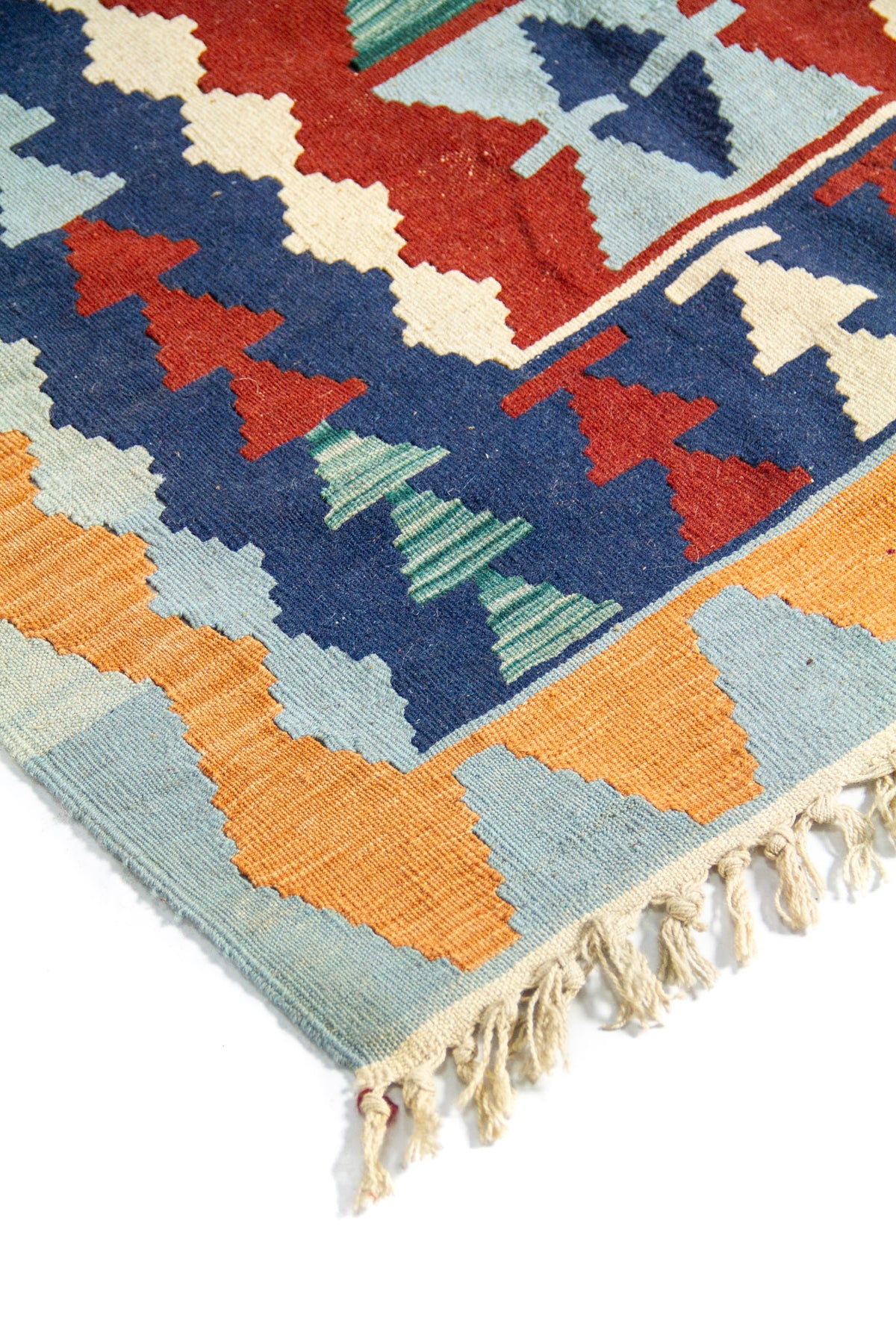 Detalle de esquina de alfombra con borde celeste y naranja, figuras geométricas en azul, rojo y verde, y flecos tejidos en tono natural.