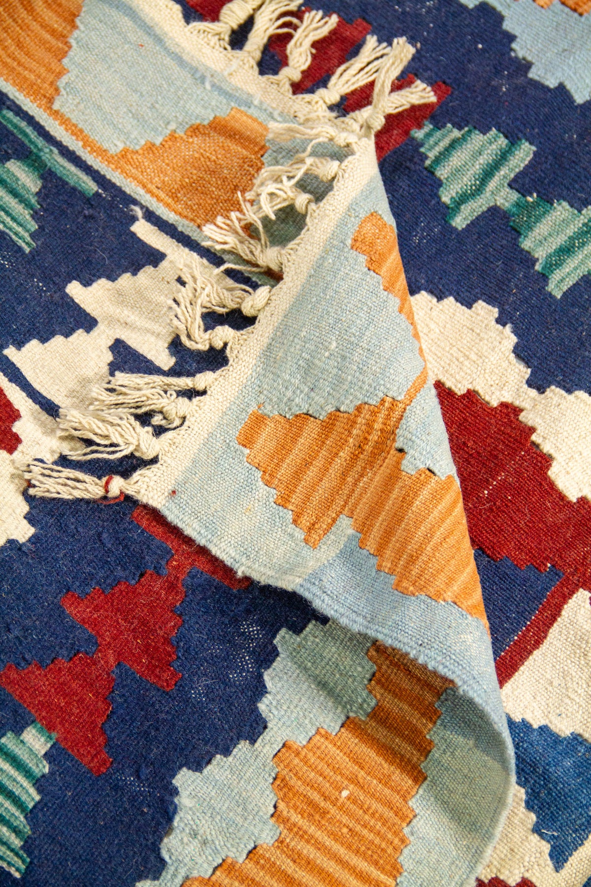 Detalle de alfombra doblada que muestra el reverso, los flecos y los motivos geométricos en tonos celeste, azul marino, rojo, verde y blanco.