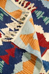 Detalle de alfombra doblada que muestra el reverso, los flecos y los motivos geométricos en tonos celeste, azul marino, rojo, verde y blanco.