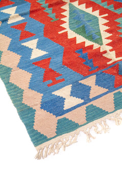 Detalle de esquina de alfombra kilim con flecos y patrones geométricos en azul, rojo y beige.