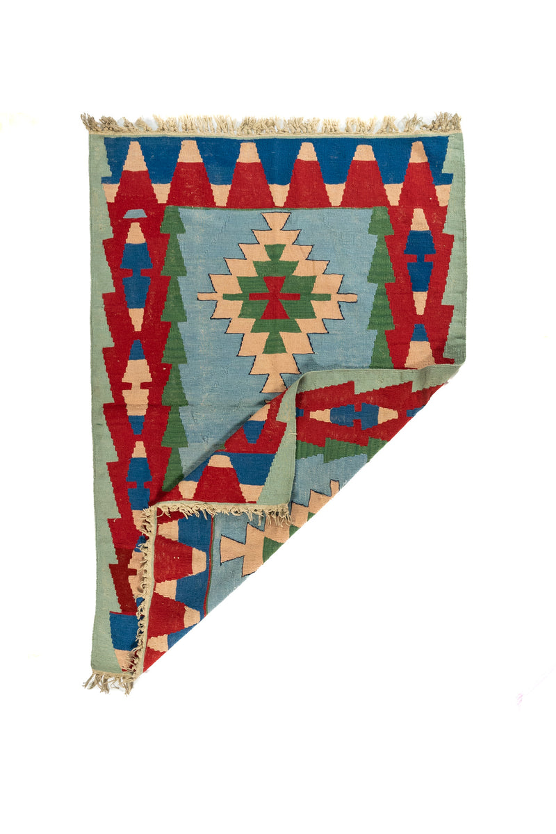 Alfombra geométrica tipo kilim con una esquina doblada que deja ver el reverso y su tejido artesanal.