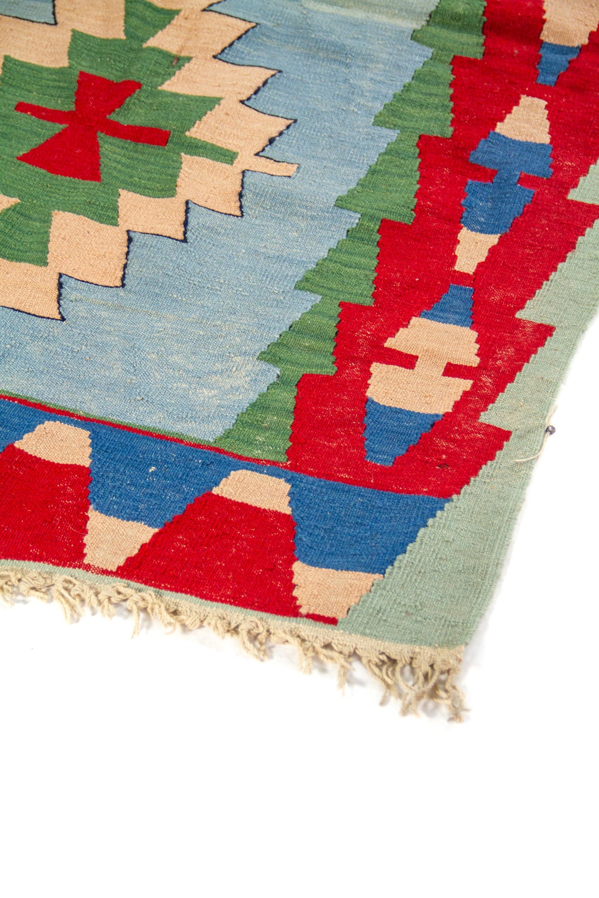 Detalle de esquina de alfombra kilim con flecos y patrones geométricos en rojo, azul, verde y beige.