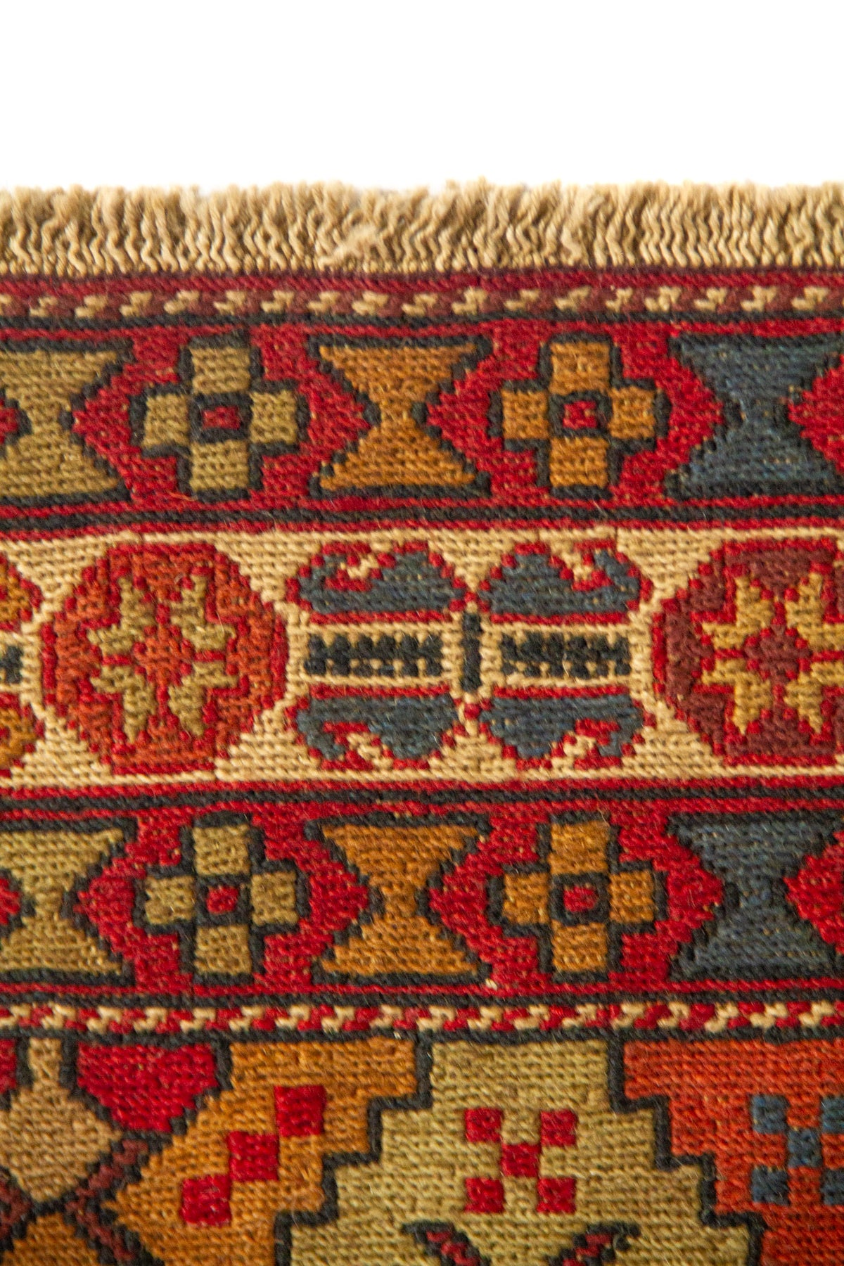 Detalle de borde de alfombra tejida a mano con pequeño fleco y motivos geométricos en rojo, beige, dorado y azul