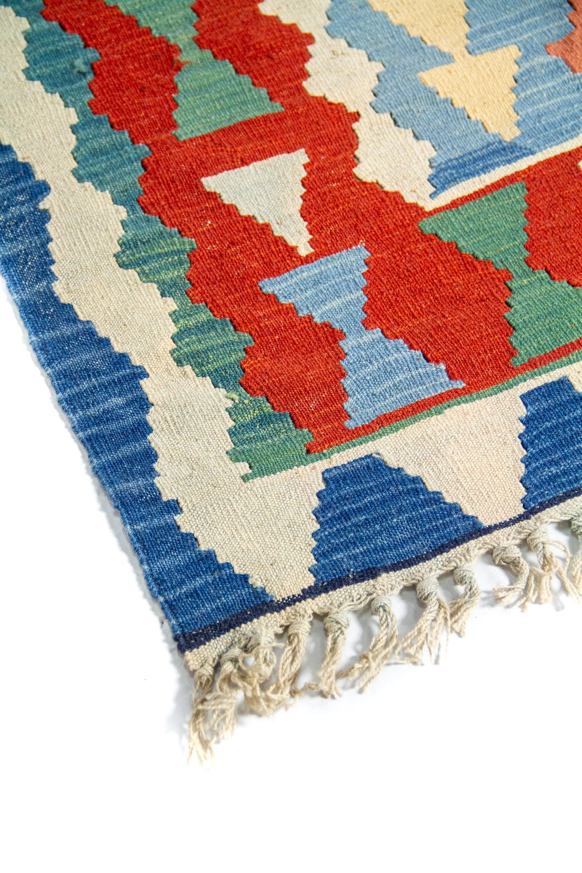 Detalle de esquina de alfombra artesanal con borde azul, motivos geométricos en rojo y celeste, y flecos color blanco suave