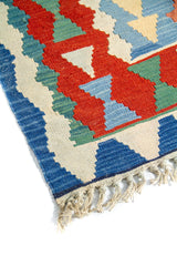 Detalle de esquina de alfombra artesanal con borde azul, motivos geométricos en rojo y celeste, y flecos color blanco suave