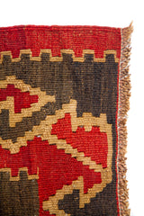 Primer plano del borde de la alfombra con flecos laterales y tejido geométrico en rojo, beige y negro.