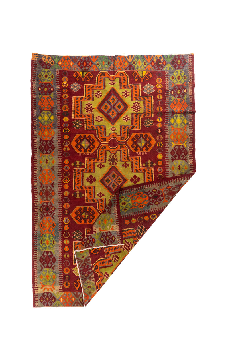 Alfombra geométrica tribal con esquina doblada que muestra su flexibilidad, en colores rojo profundo, naranja, dorado y detalles en gris 