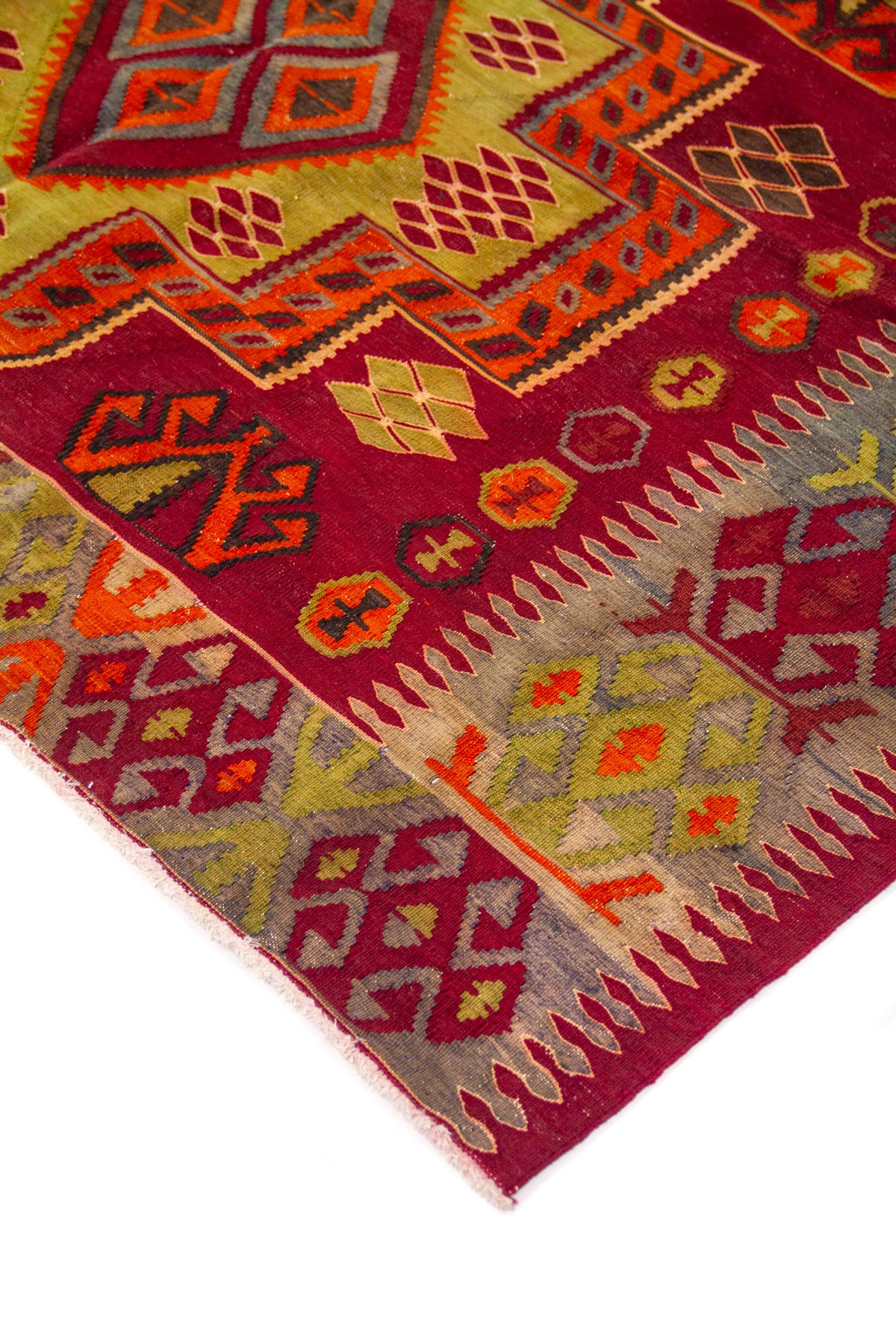Detalle de esquina de alfombra artesanal con borde geométrico en tonos gris, rojo, naranja y verde, sobre fondo rojo intenso.