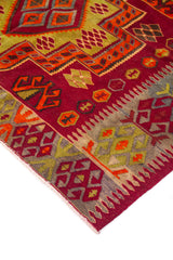 Detalle de esquina de alfombra artesanal con borde geométrico en tonos gris, rojo, naranja y verde, sobre fondo rojo intenso.