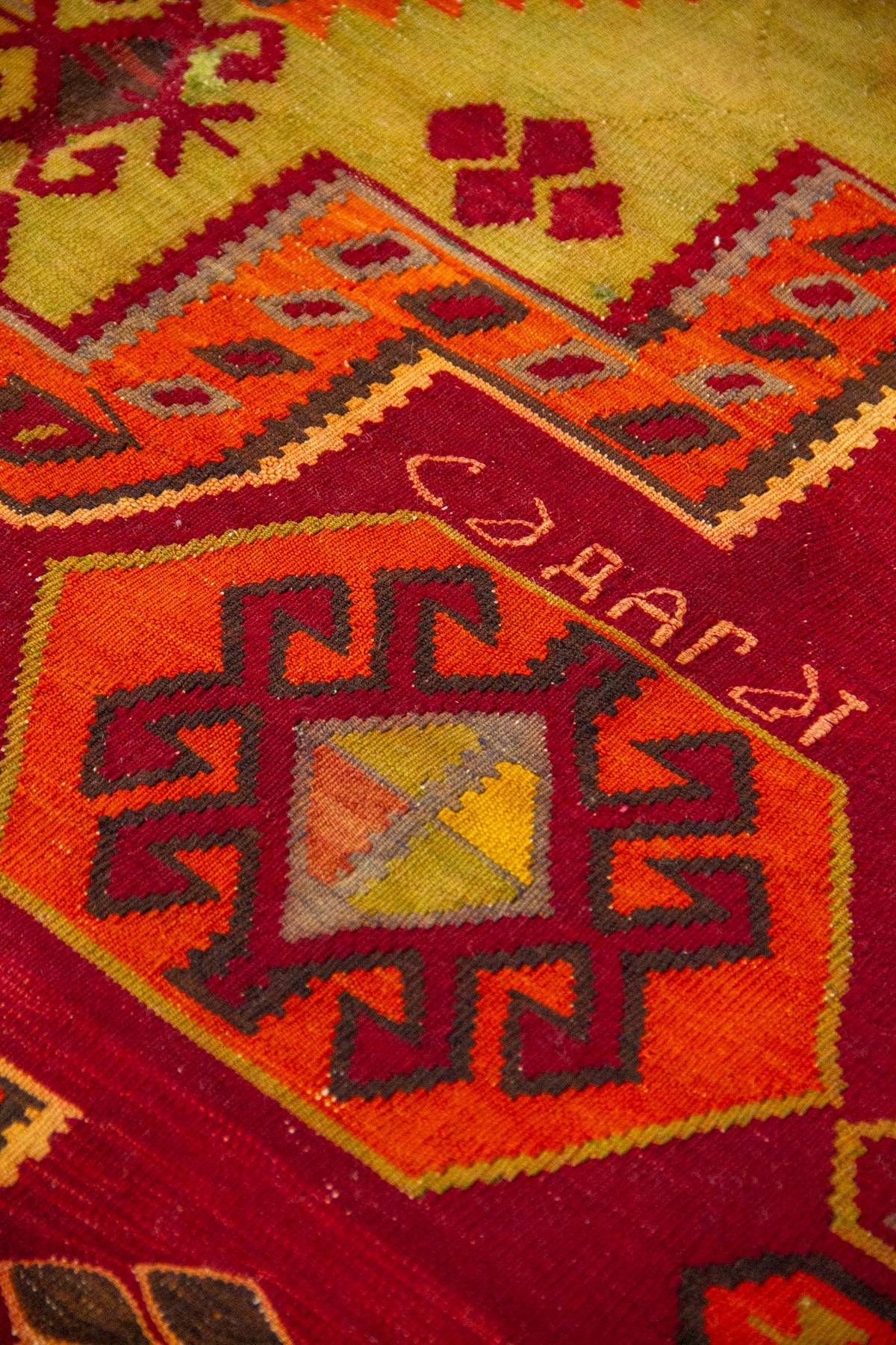 Detalle de motivo geométrico octagonal tejido en tonos naranja, rojo, amarillo y gris sobre fondo rojo.