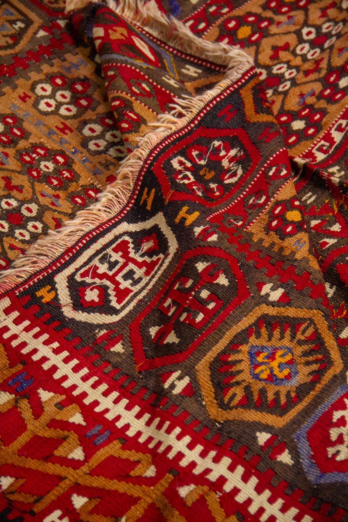 Detalle de alfombra doblada con textura artesanal, guarda geométrica y combinación de colores rojo, beige, azul y marrón