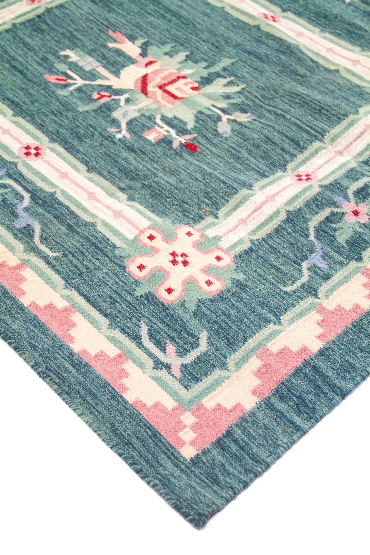 Detalle de esquina de alfombra con fondo verde y azul, líneas geométricas y acentos en rosa y blanco suave