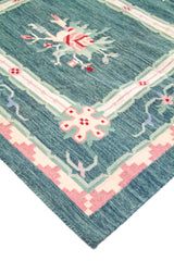 Detalle de esquina de alfombra con fondo verde y azul, líneas geométricas y acentos en rosa y blanco suave