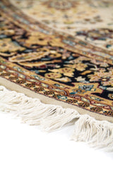 Detalle lateral de alfombra ovalada con borde negro ornamentado, flecos blancos y diseño floral tradicional