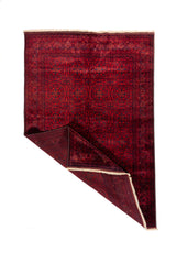 Alfombra oriental roja con una esquina doblada que muestra el reverso, diseño geométrico tribal y acabado con flecos claros.