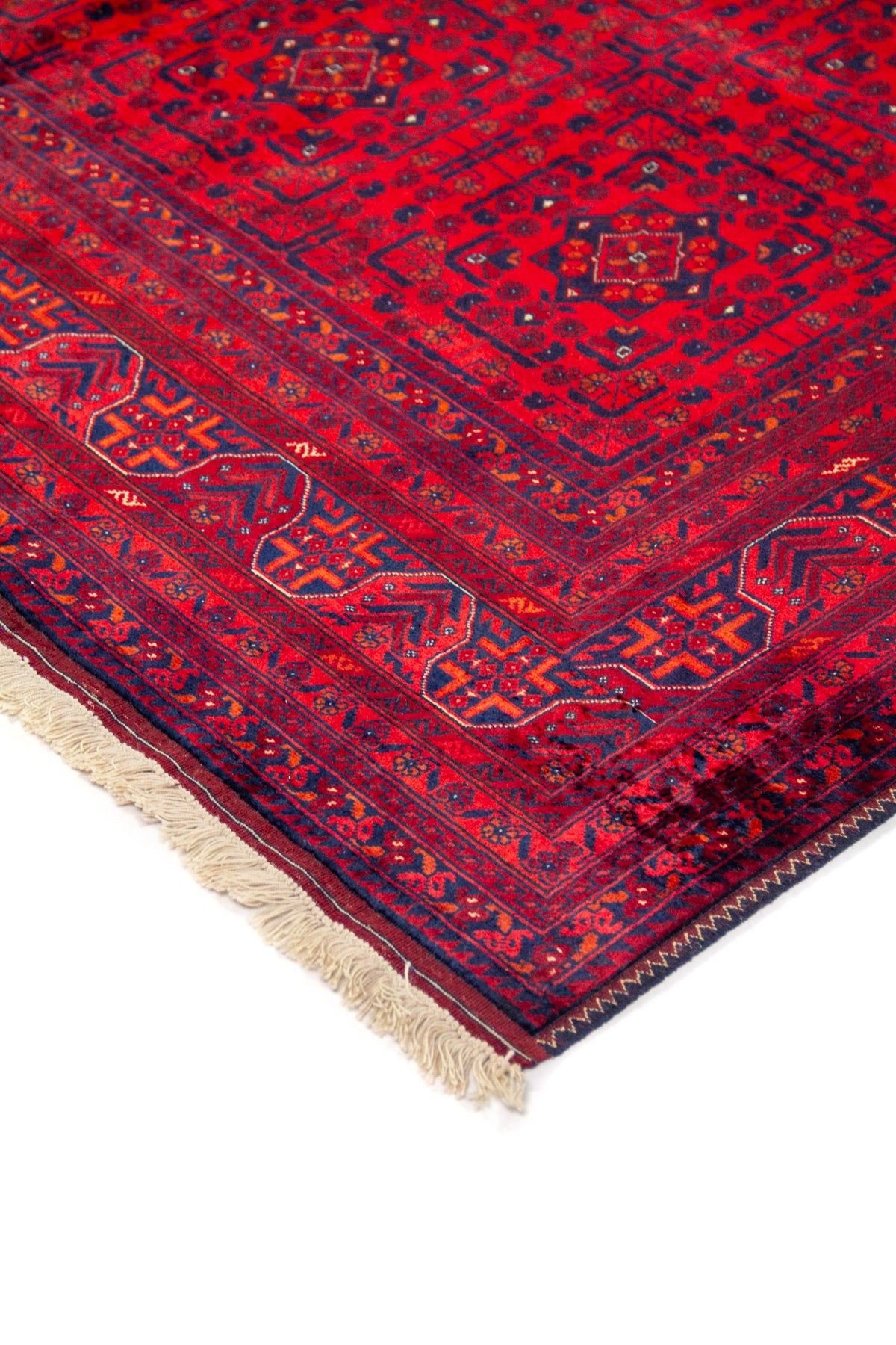 Detalle de esquina de alfombra roja con motivos geométricos en azul oscuro, borde decorativo y flecos beige.
