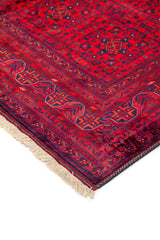 Detalle de esquina de alfombra roja con motivos geométricos en azul oscuro, borde decorativo y flecos beige.