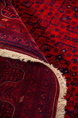 Alfombra roja enrollada mostrando la textura, el reverso tejido y los motivos geométricos tradicionales en rojo y azul oscuro.