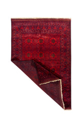 Alfombra oriental roja con esquina doblada que deja ver el reverso, detalles geométricos tradicionales y acabado con flecos beige.