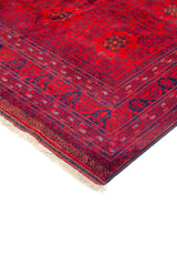 Detalle de esquina de alfombra roja con flecos claros, guarda geométrica en azul oscuro y finos patrones tribales en el borde.
