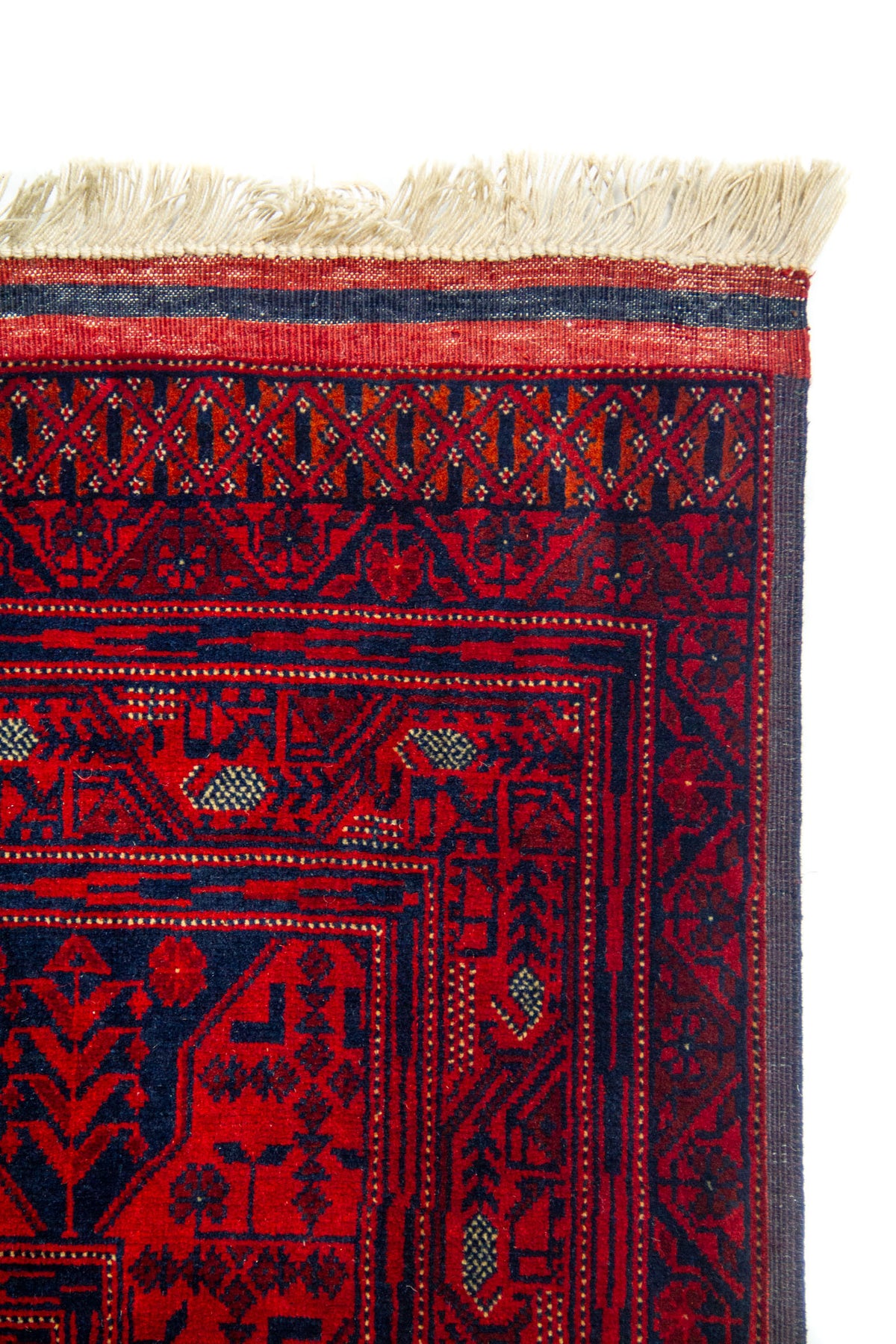 Detalle de esquina superior de alfombra roja con flecos beige y patrones geométricos tradicionales enmarcados por varias guardas decorativas.