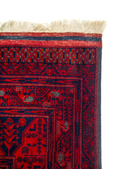 Detalle de esquina superior de alfombra roja con flecos beige y patrones geométricos tradicionales enmarcados por varias guardas decorativas.
