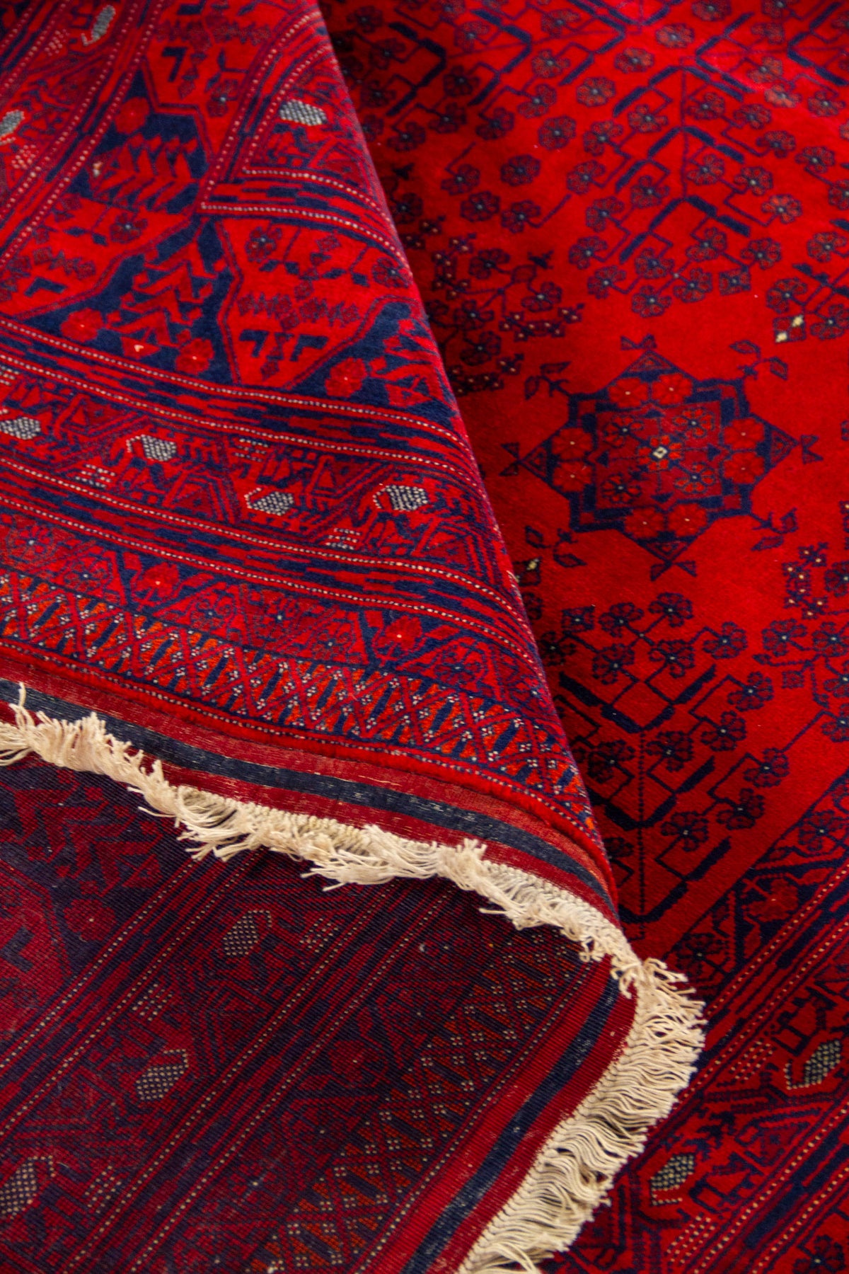 Alfombra roja parcialmente enrollada que muestra la textura, el reverso tejido y los motivos geométricos en rojo, azul oscuro y pequeños acentos claros.