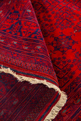 Alfombra roja parcialmente enrollada que muestra la textura, el reverso tejido y los motivos geométricos en rojo, azul oscuro y pequeños acentos claros.