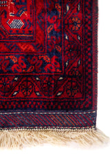 Primer plano del borde de una alfombra con diseño geométrico en rojo y azul oscuro, mostrando la textura del tejido y el acabado de la esquina.