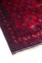 Vista en ángulo de alfombra tradicional con fondo rojo profundo, medallones geométricos repetidos y guarda decorativa de inspiración tribal.