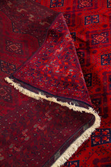 Detalle de alfombra doblada que muestra el reverso, los flecos y la estructura del tejido en una composición de tonos rojos y azul marino.