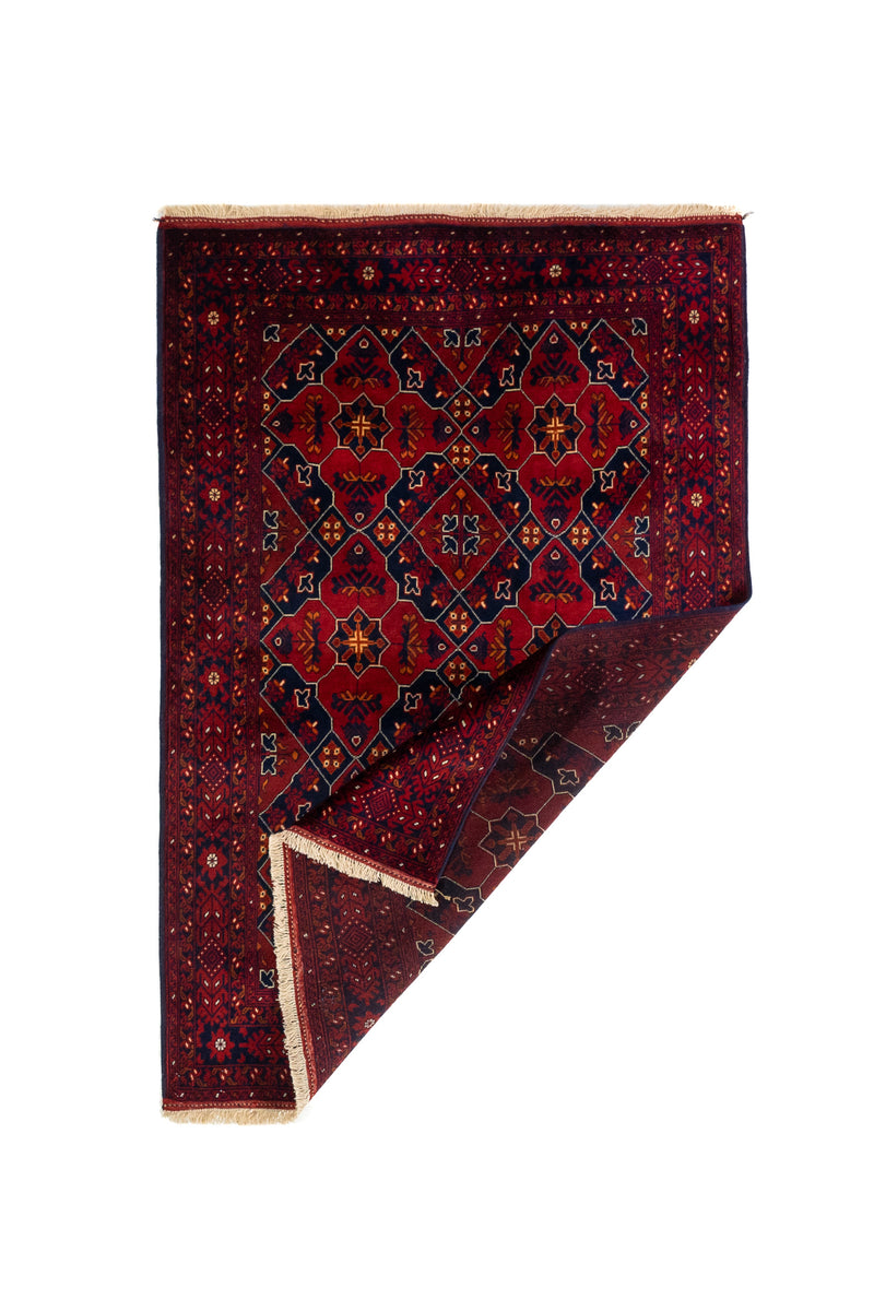 Alfombra geométrica tradicional con una esquina doblada que deja ver el reverso, el grosor del tejido y los flecos en tono beige.