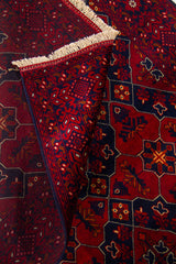 Detalle de alfombra doblada mostrando la textura del tejido, el reverso y los flecos, con diseño geométrico en rojo y azul oscuro.
