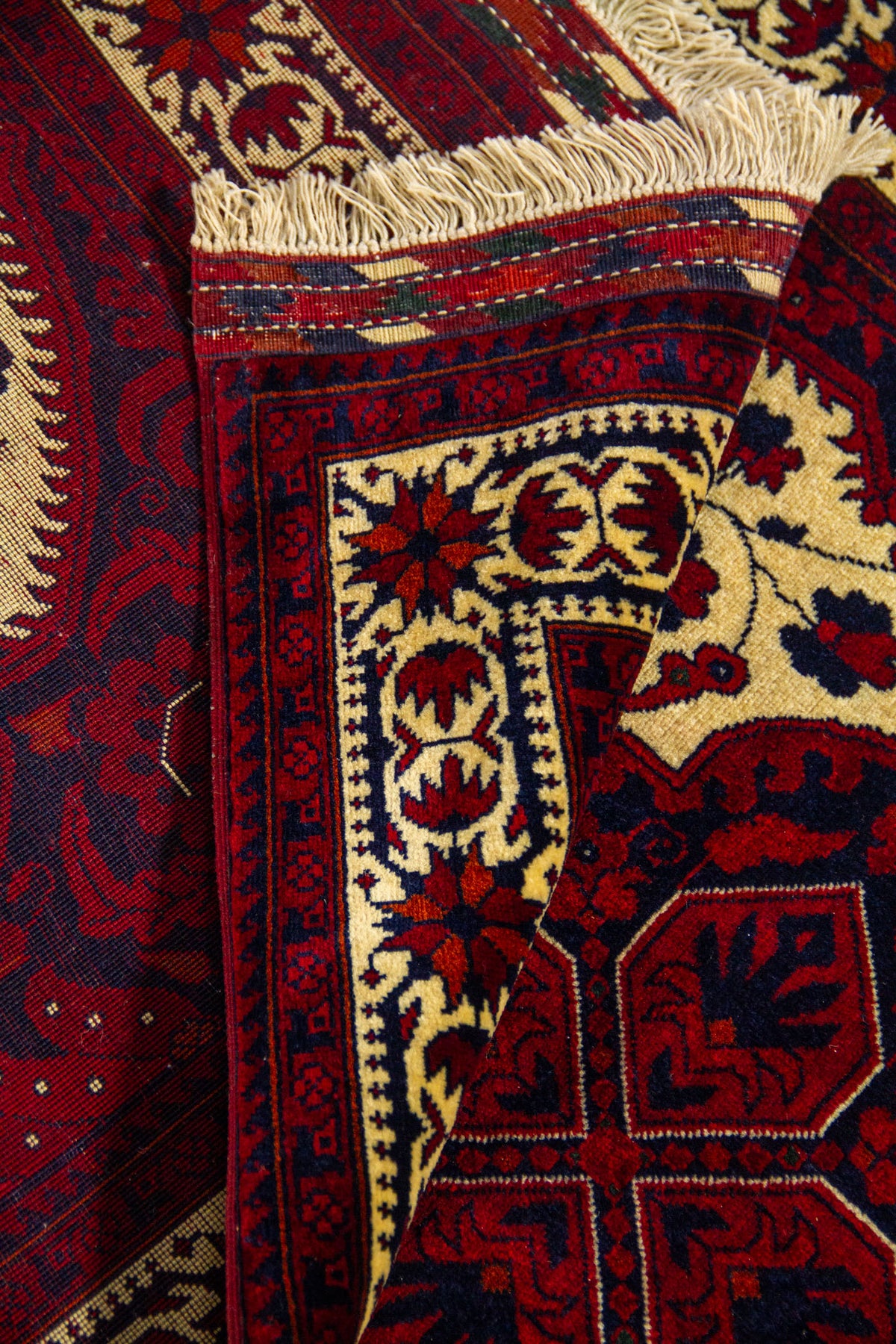 Detalle de alfombra doblada mostrando la textura del tejido, el reverso, los flecos y el contraste entre el campo central amarillo claro y los bordes rojos.