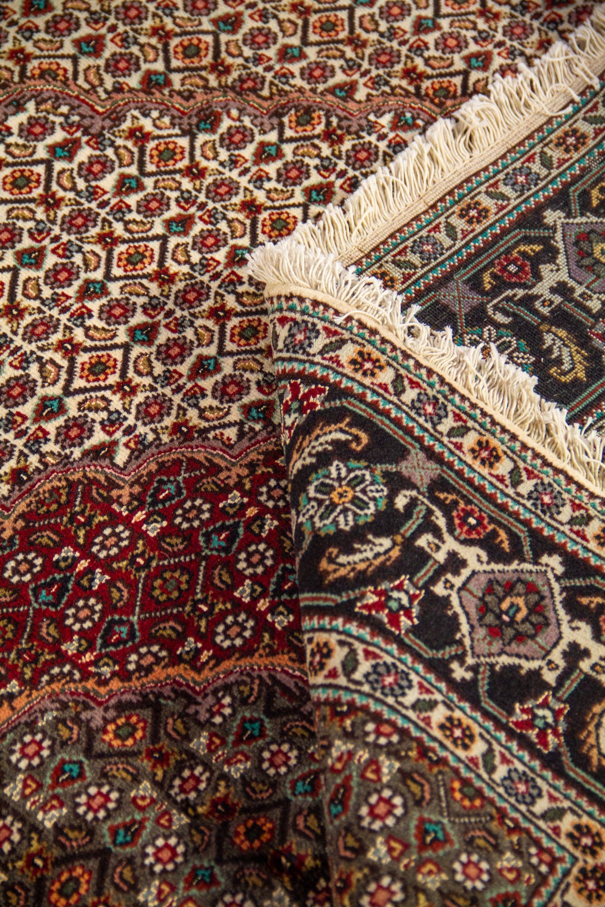 Detalle de alfombra doblada mostrando textura, flecos y diseño central en tonos rojo oscuro, blanco suave, negro y gris